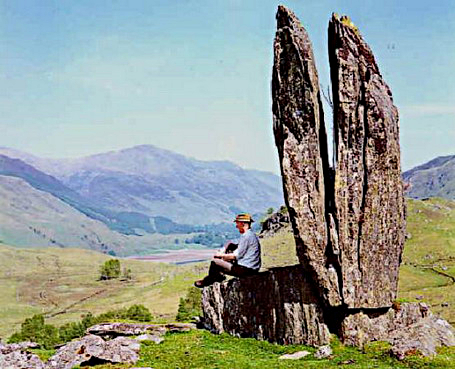 Person wearing a hat sits on a large split rock formation in a grassy valley with distant mountains and a lake in the background.